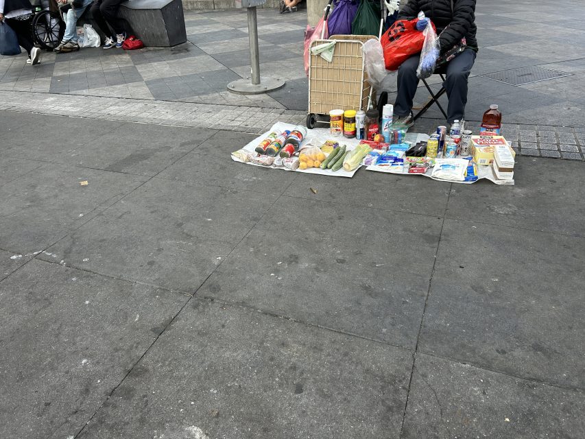 Two vendors sit on a sidewalk behind a display of assorted food items and household goods laid out on mats; a shopping cart is beside them.