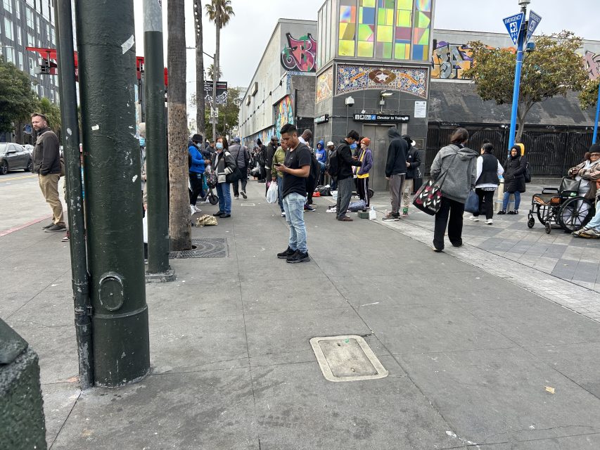 People stand and walk along a city sidewalk near buildings with colorful murals. Some are gathered in groups, and one person sits in a wheelchair.