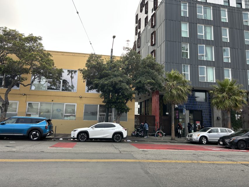Street view of parked cars, people walking on the sidewalk, and two buildings—one yellow, one gray—on an overcast day. Trees partially obscure the building fronts.