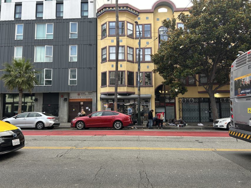 A city street with parked cars, a red bus lane, people gathered near a yellow apartment building, and a bus partially visible on the right.