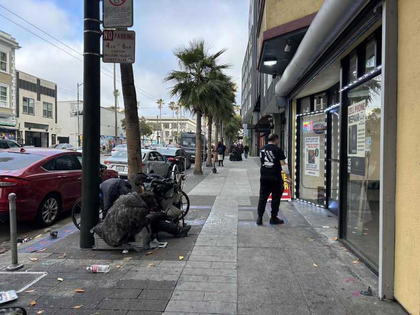A city sidewalk with parked cars, two people sitting on the ground with belongings, a man standing near a store entrance, and palm trees lining the street.