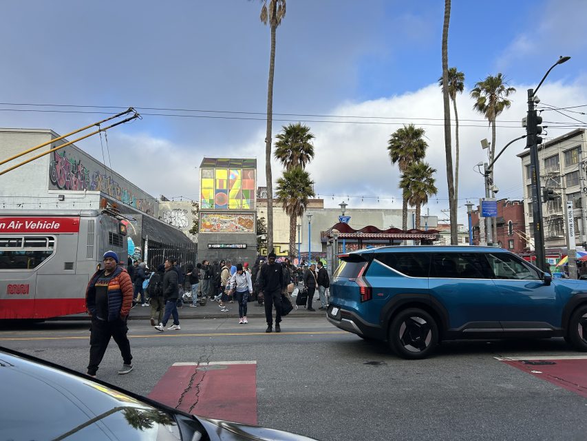 A busy city street scene with pedestrians crossing, a blue SUV and red bus stopped, palm trees, and buildings in the background under a partly cloudy sky.