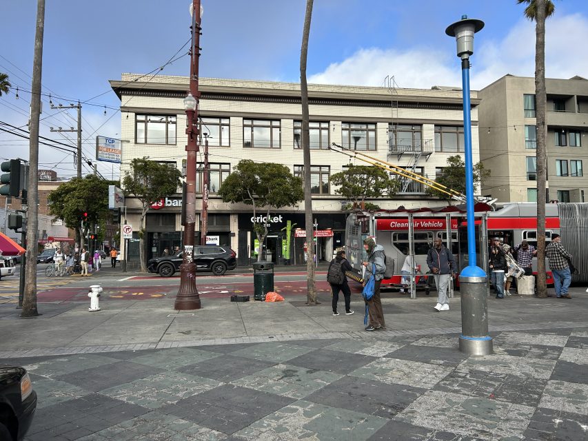 People wait near a red streetcar at an intersection in a city. Stores and parked cars line the opposite side of the street. The sky is partly cloudy.