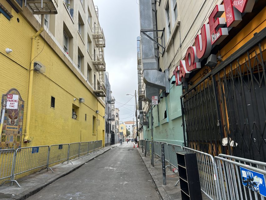 A narrow urban alleyway with metal barricades along both sides, yellow and beige buildings, and a closed storefront with a partial "TAQUERIA" sign.