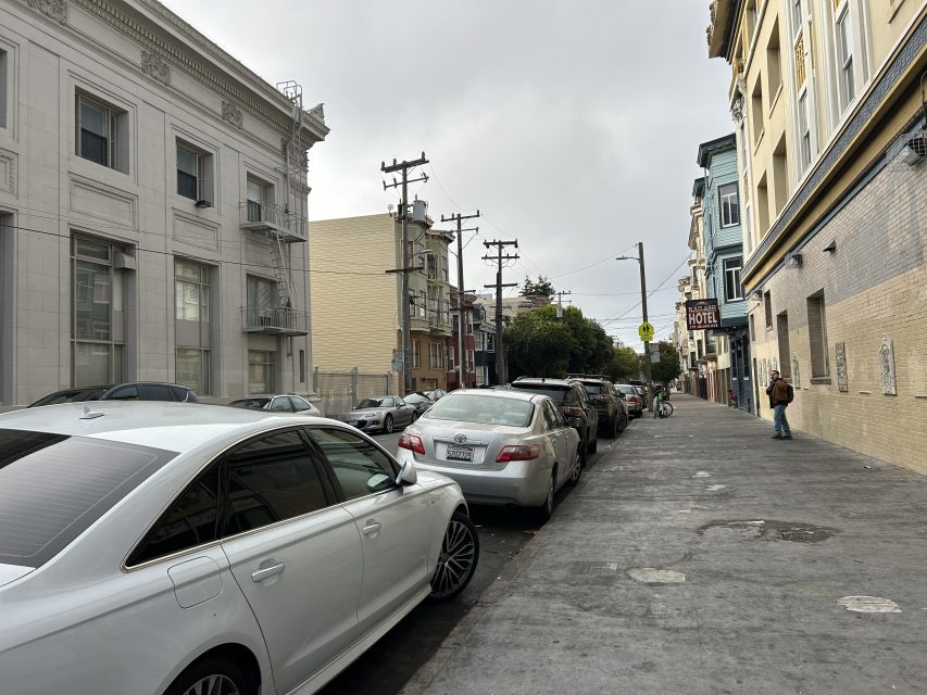 A city street with parked cars along the curb, a few pedestrians on the sidewalk, and buildings lining both sides under a cloudy sky.