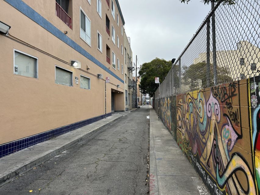 A narrow urban alleyway with graffiti on a chain-link fence on the right and beige apartment buildings on the left under a cloudy sky.