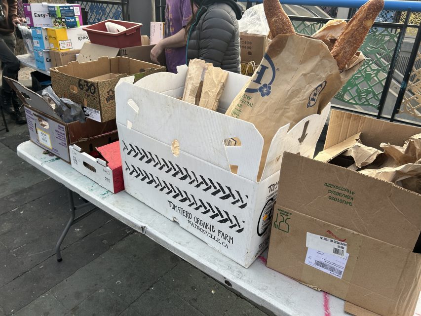 A table covered with cardboard boxes and paper bags, some containing loaves of bread, at an outdoor market or distribution event.