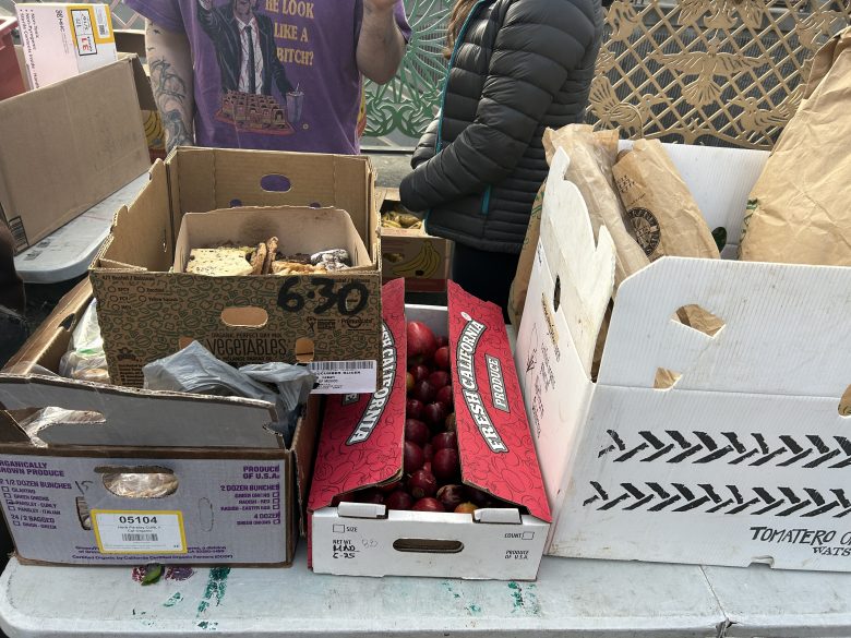 A table displays boxes of assorted produce, including radishes and vegetables; two people stand behind the table, partially visible.