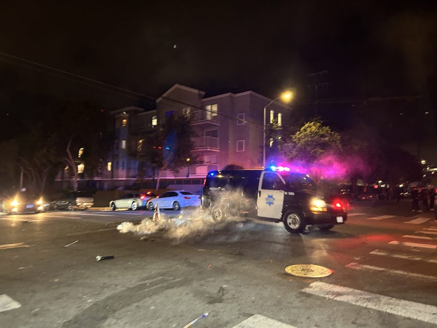 A police van with flashing lights is parked on a city street at night, with smoke rising from the ground and debris scattered nearby.