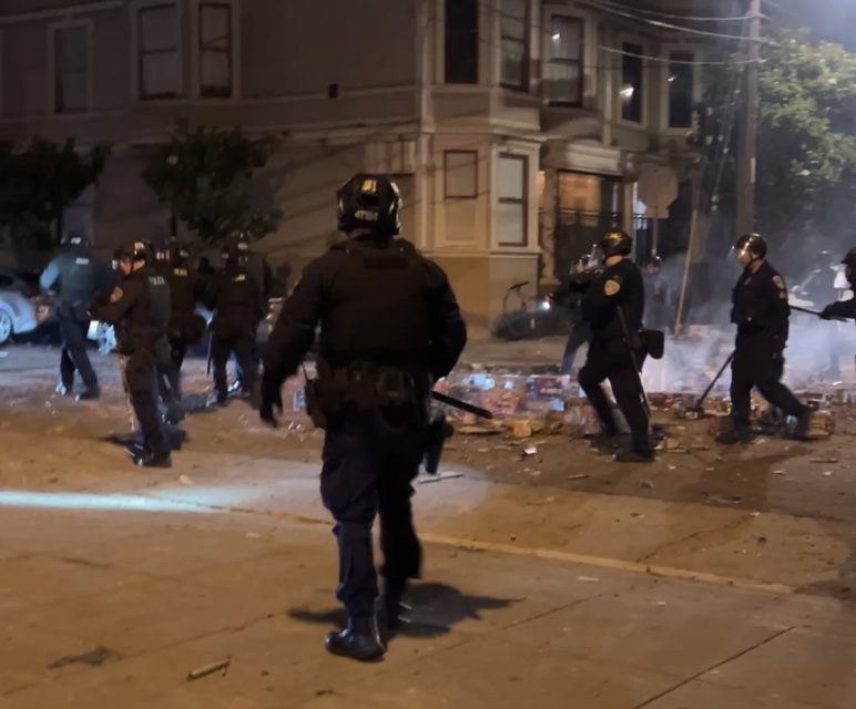 Police officers in riot gear stand on a city street at night amid debris and smoke, with a building and scattered objects visible in the background.