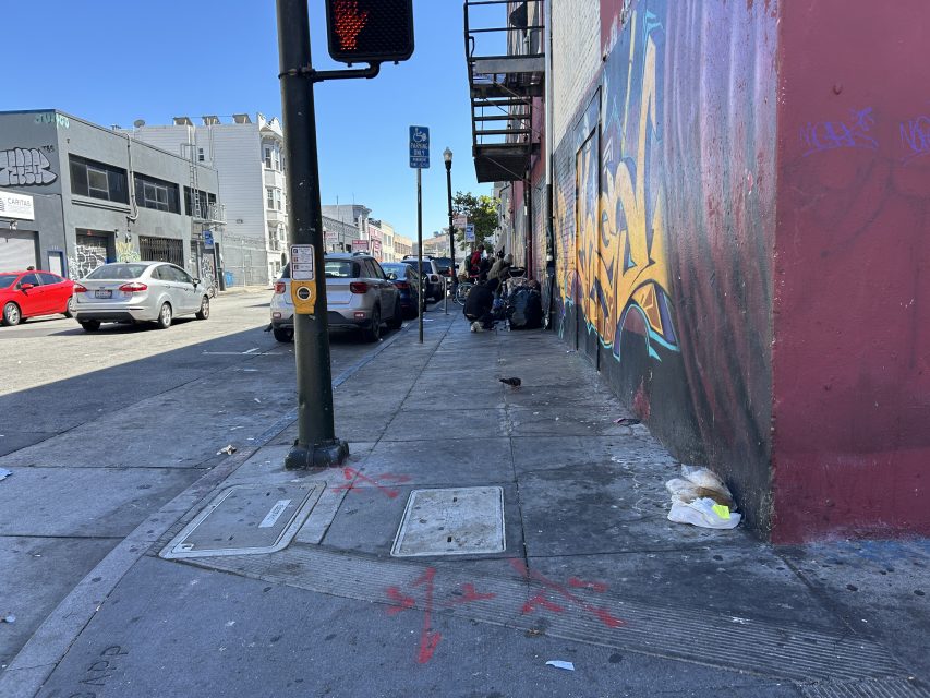 A city sidewalk with cars parked along the street, a group of people sitting by a building with graffiti, and scattered trash on the ground.