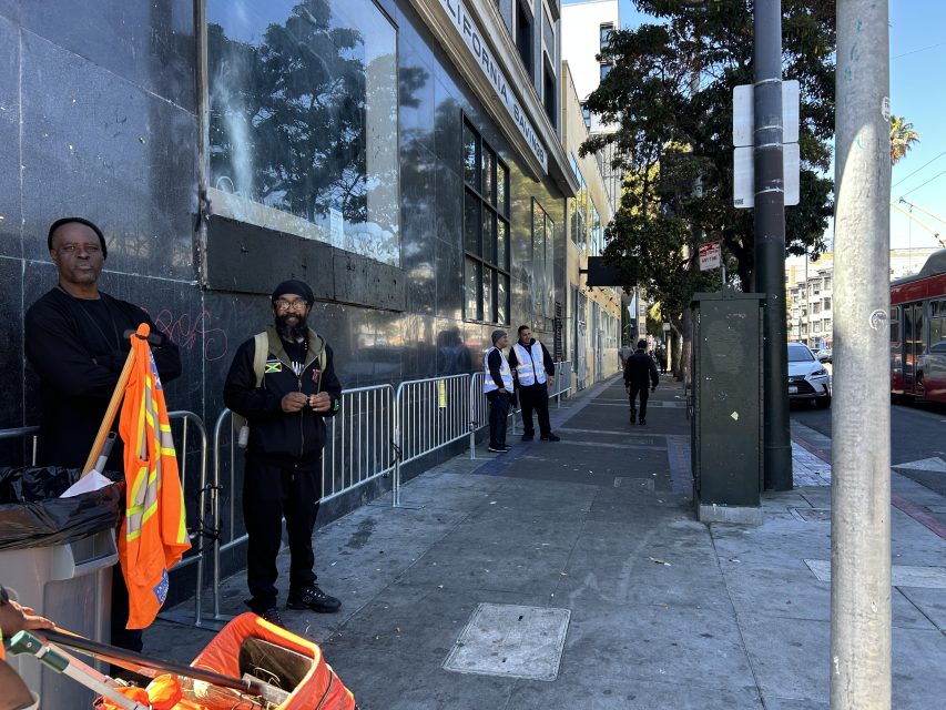 Several people stand on a city sidewalk near a building, some wearing uniforms and safety vests. A trash can with an orange vest draped over it is in the foreground.