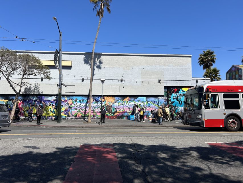 A city street with a colorful graffiti mural on a building, people gathered along the sidewalk, and a red city bus passing by under a clear blue sky.