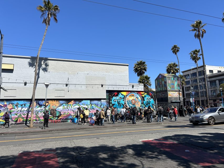 A group of people stand and walk along a city sidewalk in front of a colorful mural and buildings, with palm trees and clear blue sky overhead.