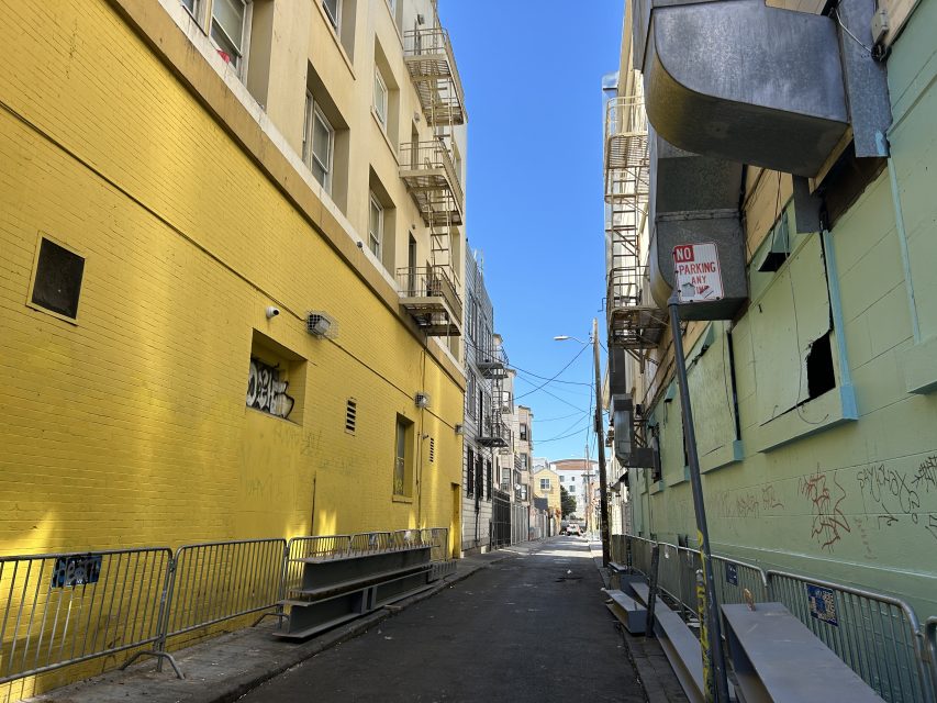 A narrow urban alleyway with yellow and green walls, metal railings, graffiti, barred windows, and a "No Parking" sign under a clear blue sky.