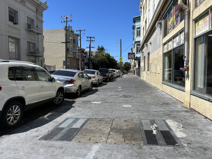 A city sidewalk lined with parked cars beside apartment buildings and a storefront on a sunny day.