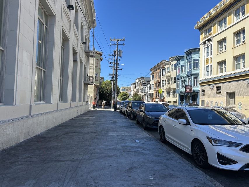 A city street with parked cars along the curb, multi-story buildings on both sides, and a clear blue sky overhead.