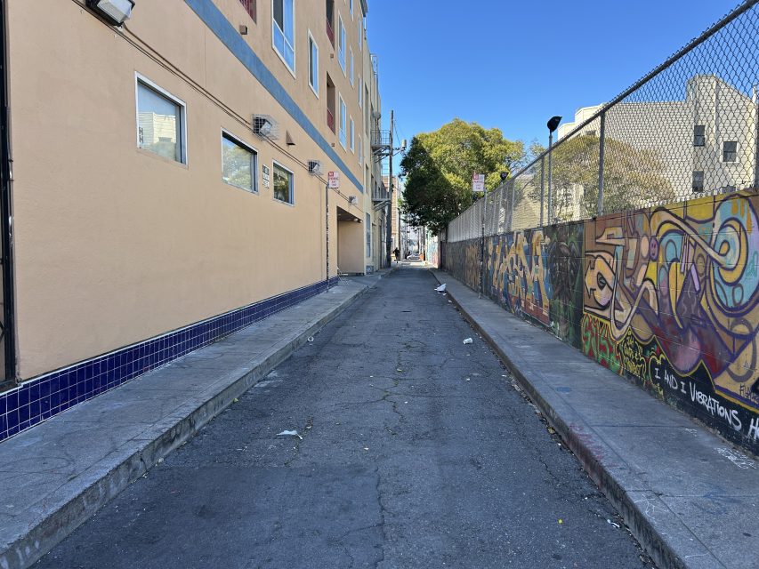 Narrow urban alley with cracked pavement, graffiti-covered wall on the right, beige building on the left, and scattered litter on the ground.