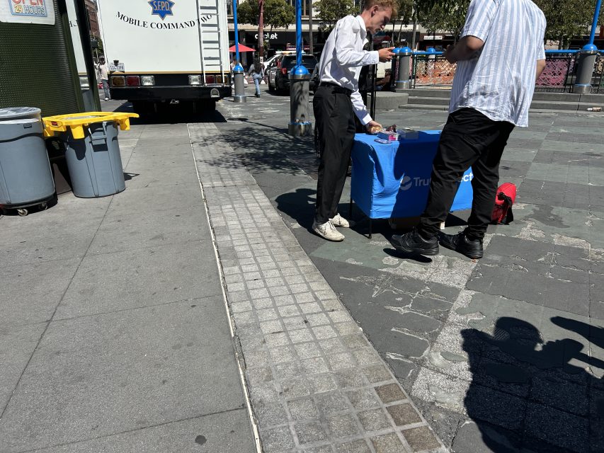 Two people stand by a blue table with pamphlets on a city sidewalk near a truck and trash bins on a sunny day.