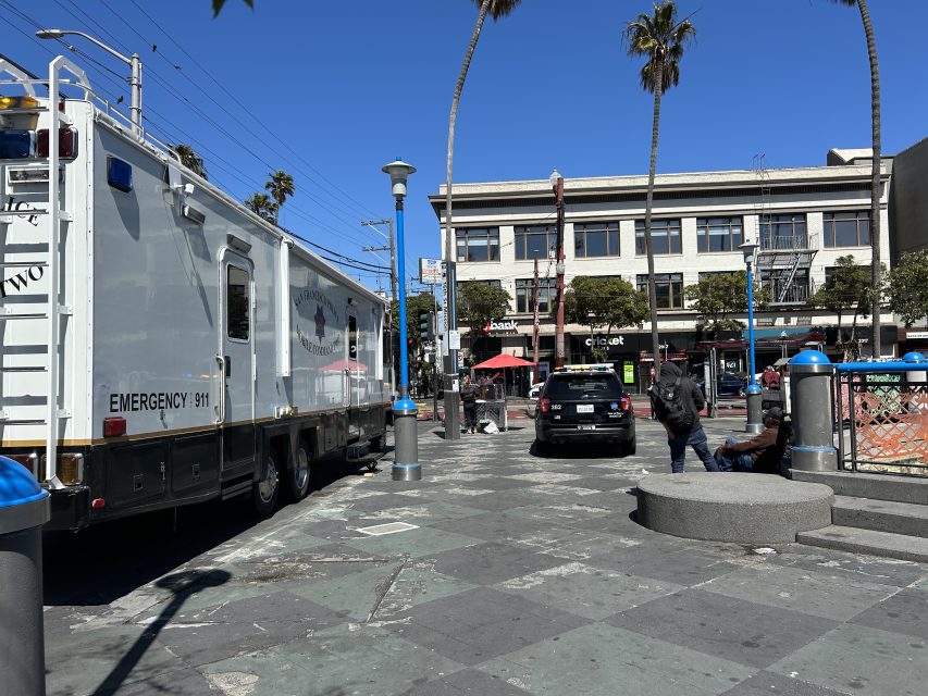 A police emergency vehicle and a police car are parked on a city plaza near people sitting on a circular bench, with shops and palm trees in the background.