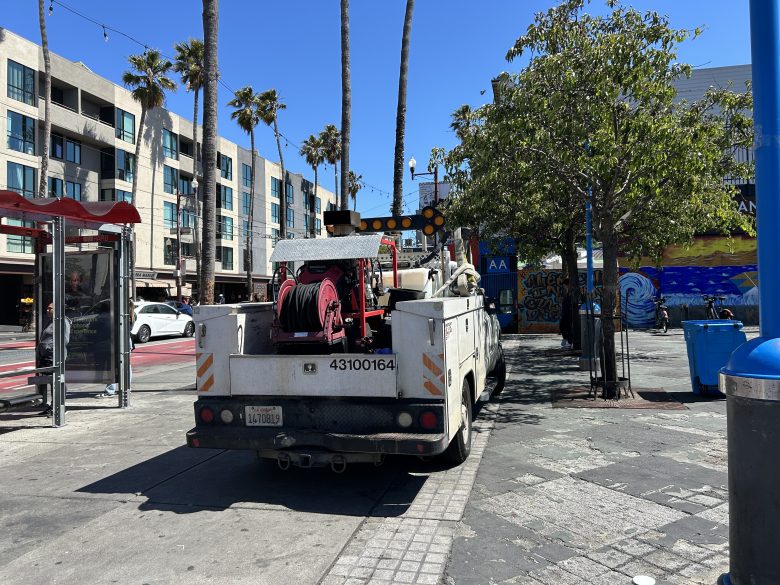 A utility truck with cables and equipment is parked on a city street near buildings, palm trees, a mural, and a blue trash bin on a sunny day.