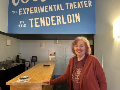 A woman with pink hair smiles while standing beside a wooden counter in a blue room with a sign reading "Experimental Theater in the Tenderloin.