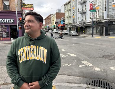 A person in a green Tenderloin hoodie stands at a city street corner, surrounded by bustling buildings, traffic lights, and passing pedestrians.