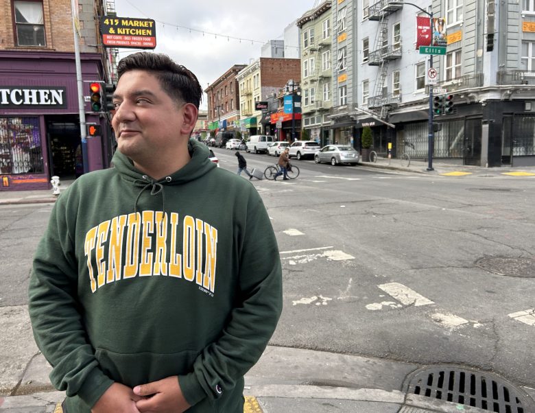 A person in a green Tenderloin hoodie stands at a city street corner, surrounded by bustling buildings, traffic lights, and passing pedestrians.