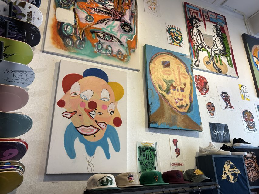 A wall displays abstract and surreal paintings along with skateboards and hats arranged on shelves below.