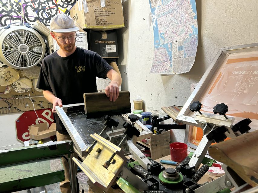 A man wearing a white hard hat and glasses operates a screen printing press in a cluttered workspace with various tools, boxes, and a map on the wall.