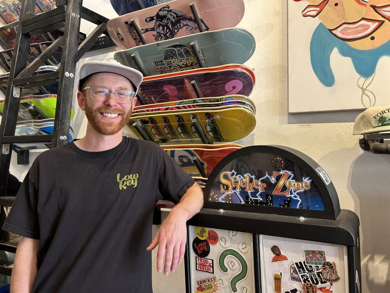 A man wearing glasses and a cap stands in a skateboard shop, smiling next to a Sticker Zone vending machine and racks of colorful skateboards.