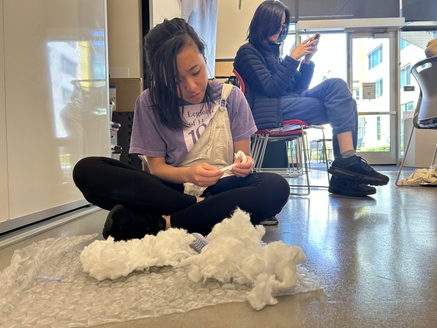 A person sits cross-legged on the floor handling white stuffing material, while another person sits on a chair in the background using a smartphone.