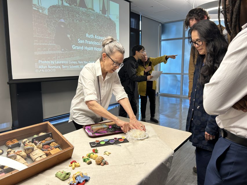 A woman arranges clay art pieces on a table as several people observe. A display case and art supplies are nearby. A presentation is projected in the background.