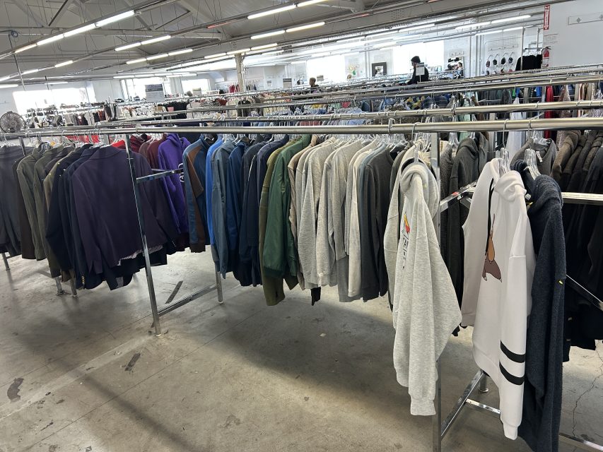 Clothing racks filled with jackets, hoodies, and sweaters are arranged in a spacious, well-lit thrift store with concrete floors.