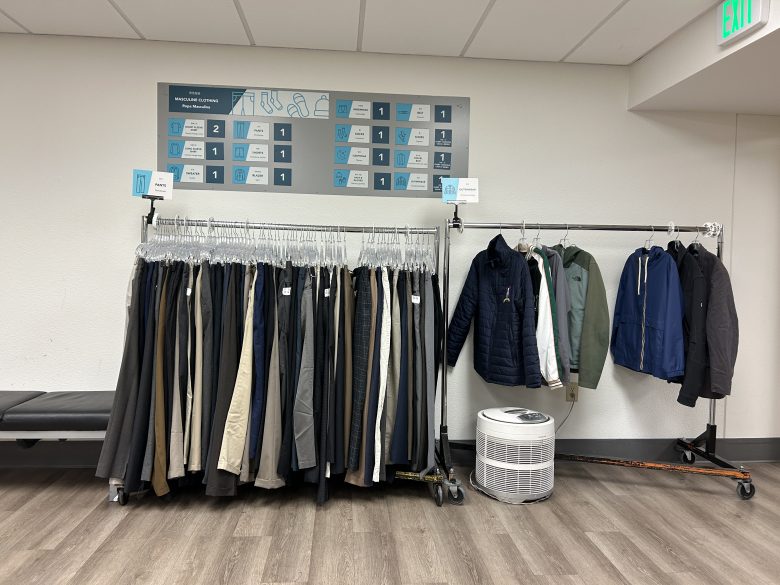 Two clothing racks hold assorted pants and jackets in an indoor room with a chart on the wall and an air purifier on the floor.