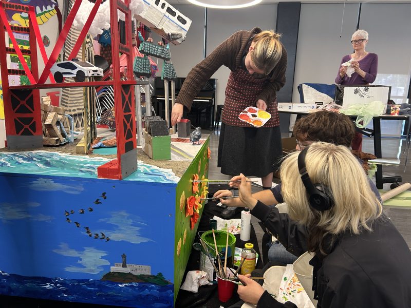 Three people paint a colorful model featuring a bridge, cars, and a seascape, while another person stands in the background. Art supplies are scattered around them.