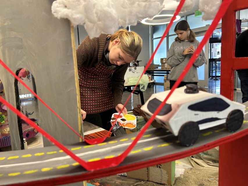 A person paints a model bridge with a toy car on it, while another person works in the background in a room with craft supplies.