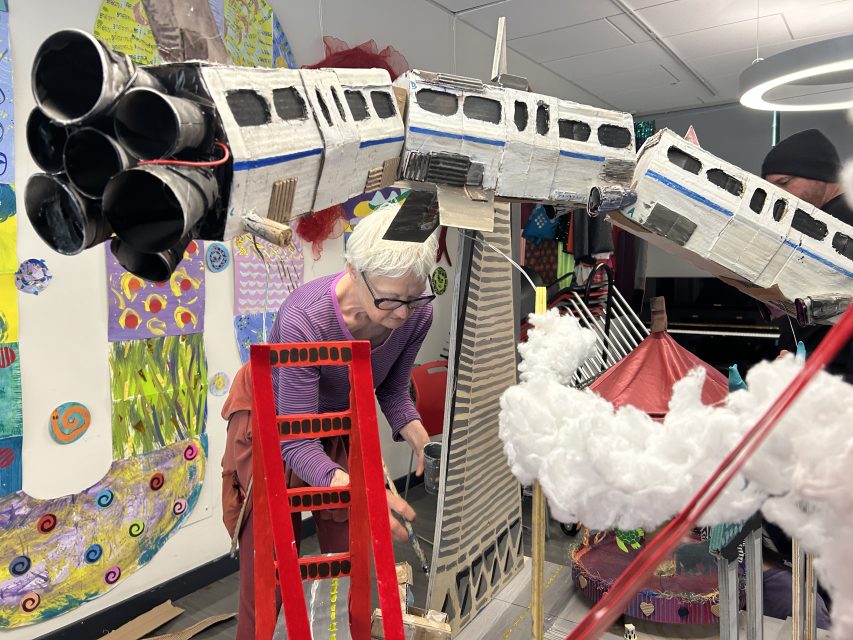 A person works on a colorful art installation featuring model trains, clouds, and various structures in a decorated indoor space.