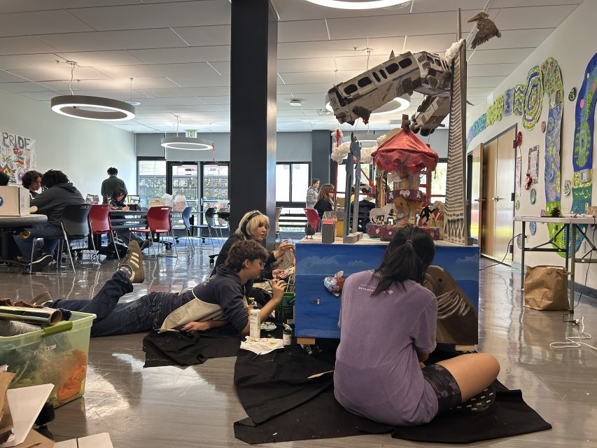 Several people work on painting and assembling colorful art projects in a spacious, well-lit indoor classroom or studio.