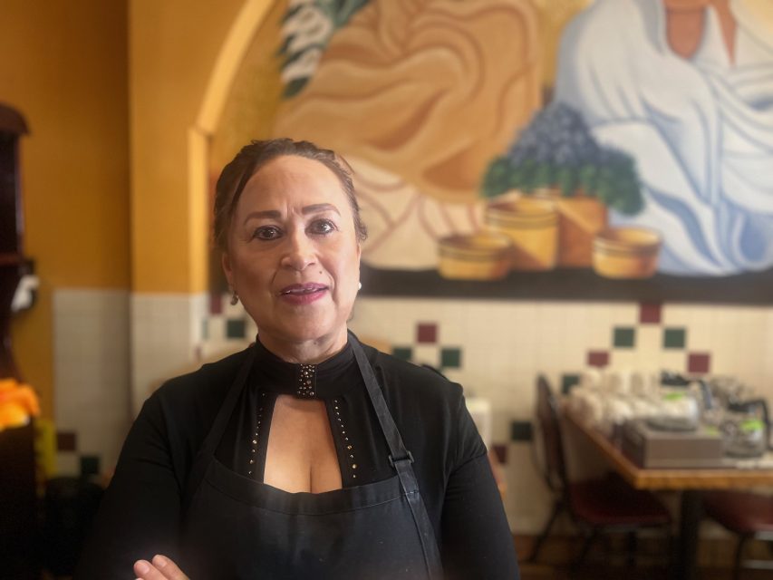 A woman in a black top and apron stands with arms crossed inside a restaurant, with a colorful mural and neatly arranged tables in the background.