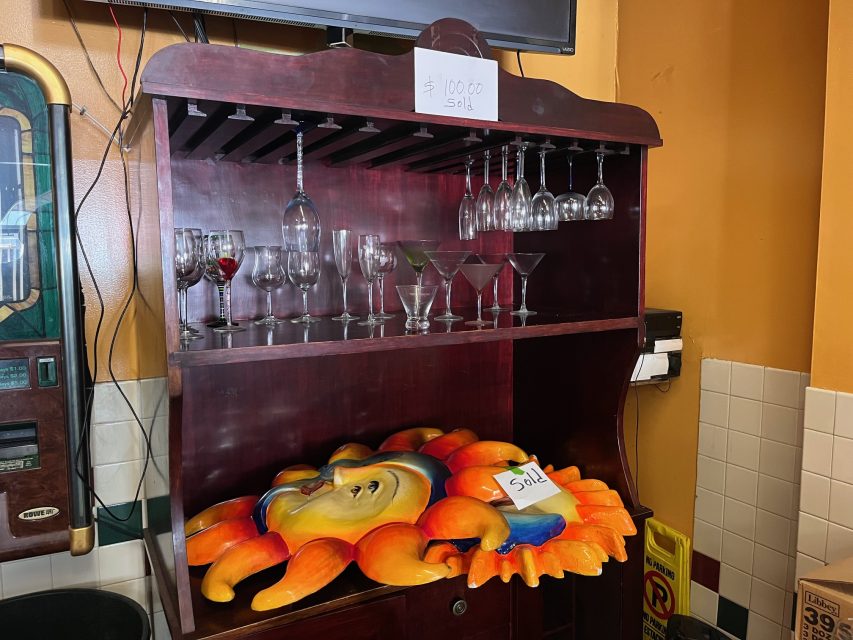 A wooden cabinet holds various wine and cocktail glasses; a decorative crab platter labeled "Sold" sits on the bottom shelf. A "Sold" sign with "$100.00" is taped to the cabinet.