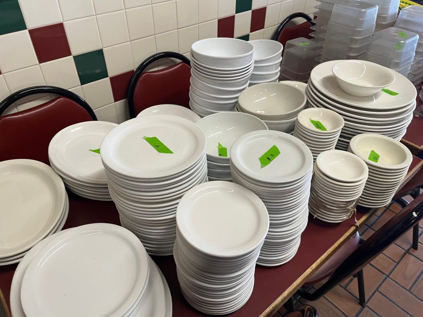 Stacks of white plates, bowls, and dishes with green price tags are arranged on tables against a tiled wall with red and green accents.