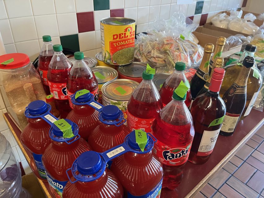 A table displays bottles of red Fanta, wine, canned goods, a large can of tomatillos, packaged food, and other assorted grocery items, each marked with a green price sticker.