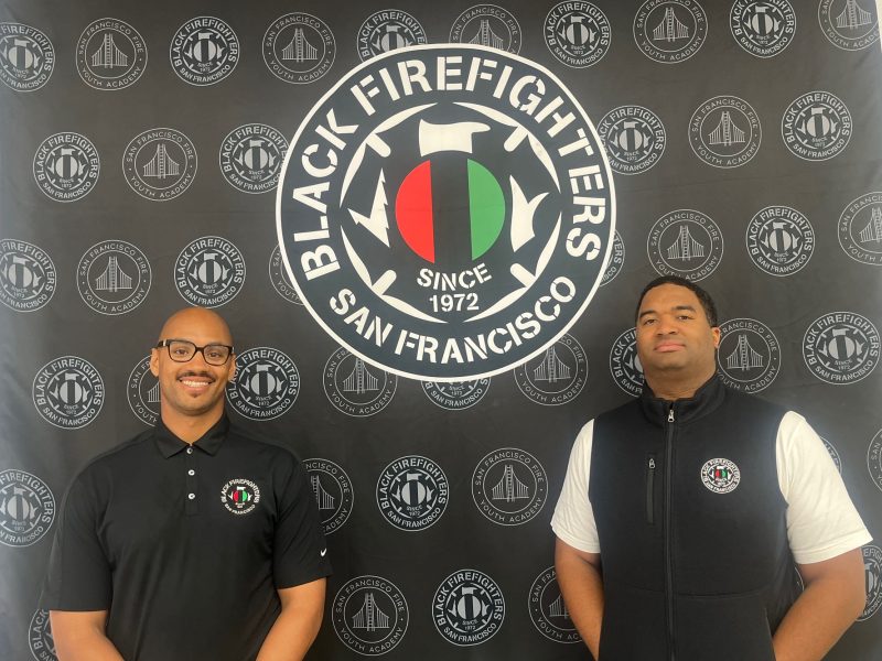 Two men stand in front of a banner that reads "Black Firefighters San Francisco Since 1972," featuring a red, black, and green emblem.