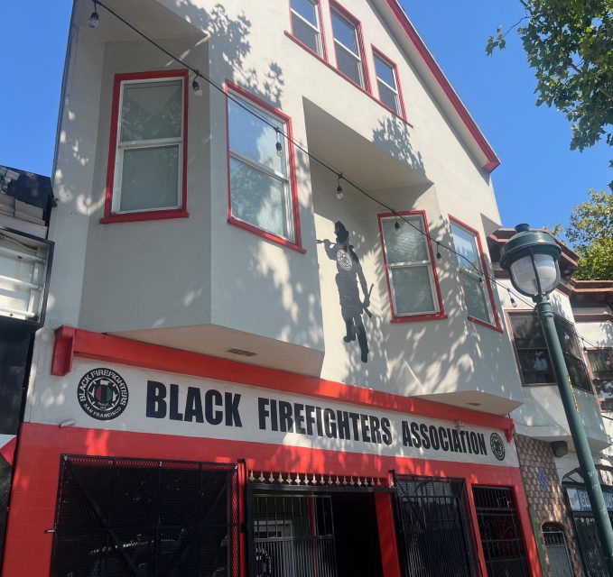 A building with a sign reading "Black Firefighters Association" on the ground floor, red and white exterior, and a firefighter silhouette on the upper facade.