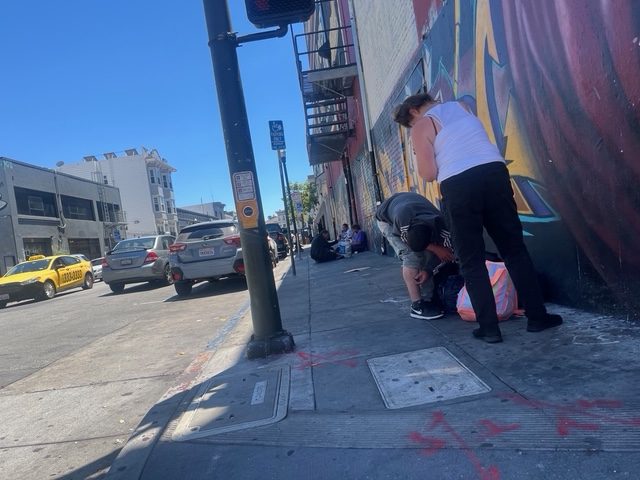 People stand and sit on a city sidewalk near a mural-covered wall. Cars and a yellow taxi are parked on the street under a sunny, clear sky.