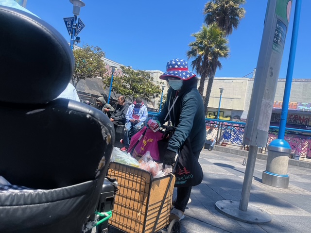 A person wearing a star-spangled hat pushes a shopping cart filled with bags in an urban outdoor setting, with people and palm trees in the background.