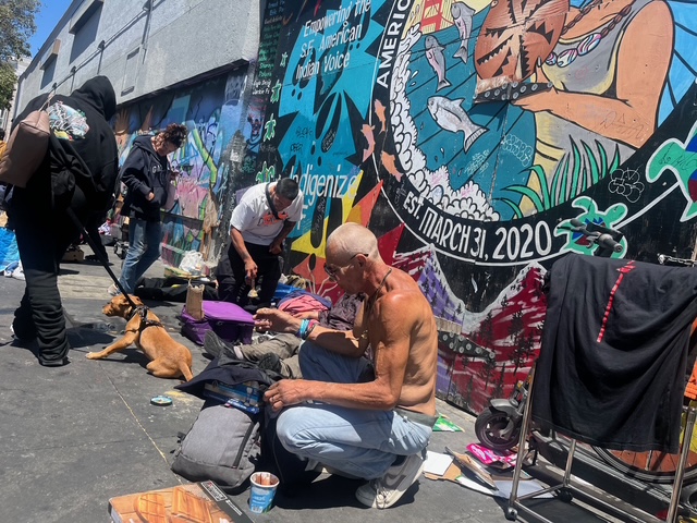 Several people gather on a sidewalk beside a mural-covered wall, with personal belongings and a dog resting nearby. One man sits shirtless in the foreground.