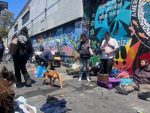 A group of people, some sitting and some standing, gather on a graffiti-covered urban street with scattered belongings and a dog in the foreground.