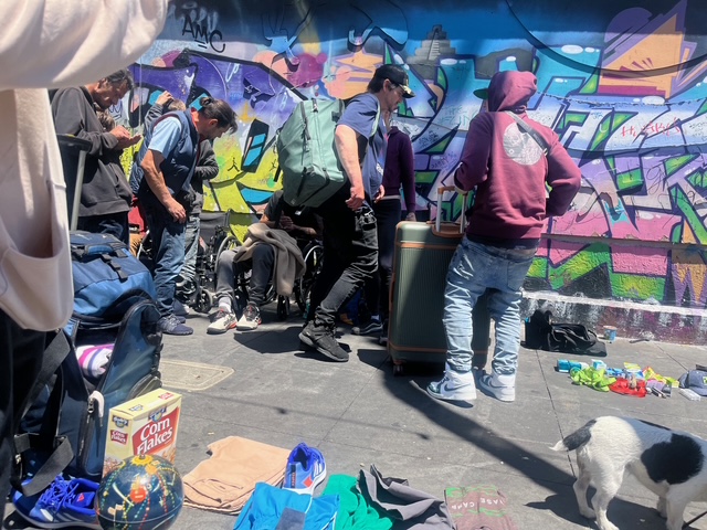 People gather on a city sidewalk with bags and belongings, near a colorful graffiti wall. Clothes and a Corn Flakes box are on the ground; a dog is in the foreground.